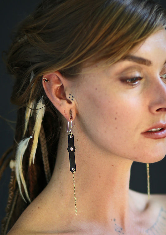 A close-up image of a woman wearing link earrings with leather hooks and a brass chain, featuring eyelets and studs.