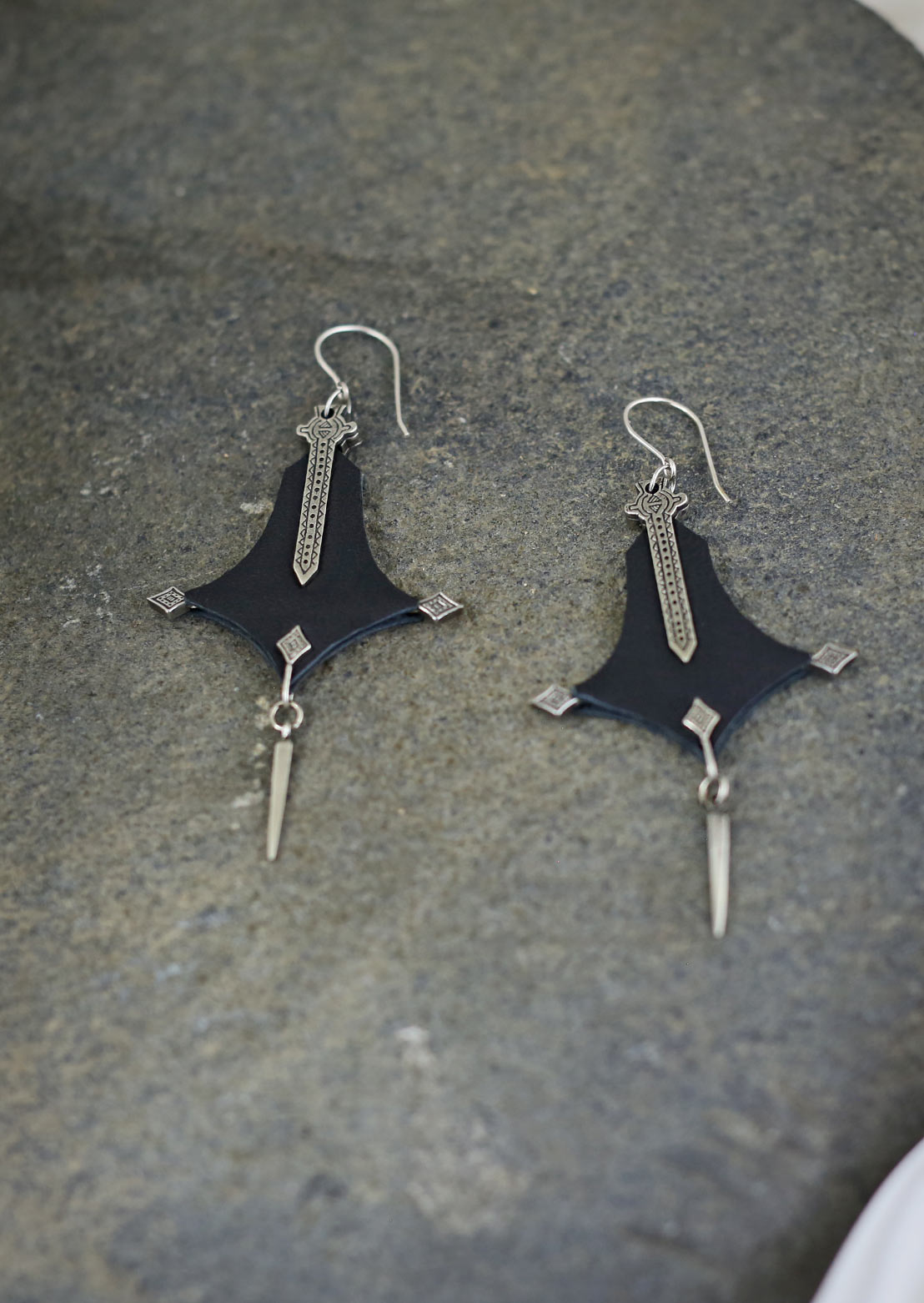 A pair of Nomad leather earrings with double leather panels and intricate brass ornamental panels, displayed on a white surface with a decorative background.
