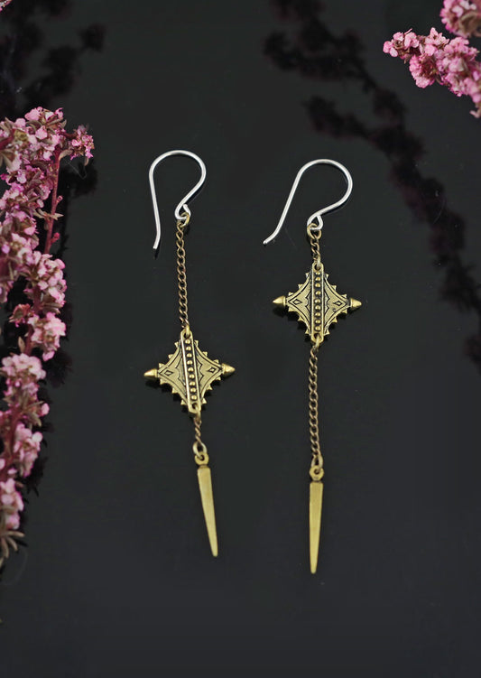 A pair of Nubian Twin Earrings with diamond-shaped brass ornaments and pendulum tips, displayed against a dark background with floral accents.