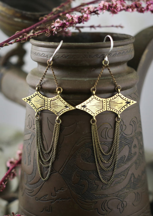 A pair of gold-colored, double spade-shaped earrings with intricate African-inspired markings, hanging from a delicate chain, displayed against a patterned ceramic background.