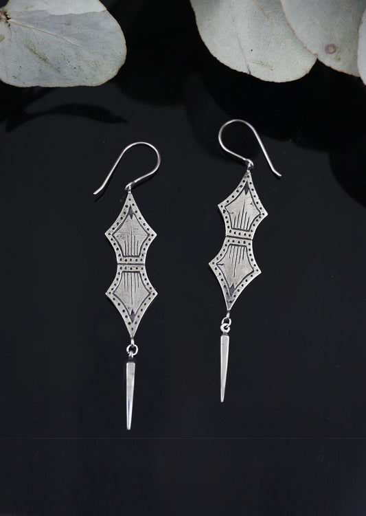 A pair of dangling earrings with a tribal geometric design, crafted from brass with sterling silver hooks, displayed against a dark background.