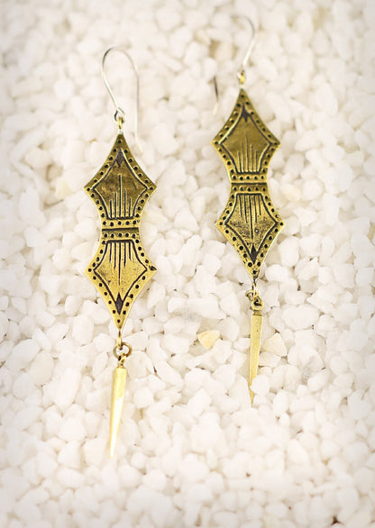 A person wearing Primal Link Earrings with a subtle tribal design, featuring double spade-shaped brass ornaments and sterling silver hooks.