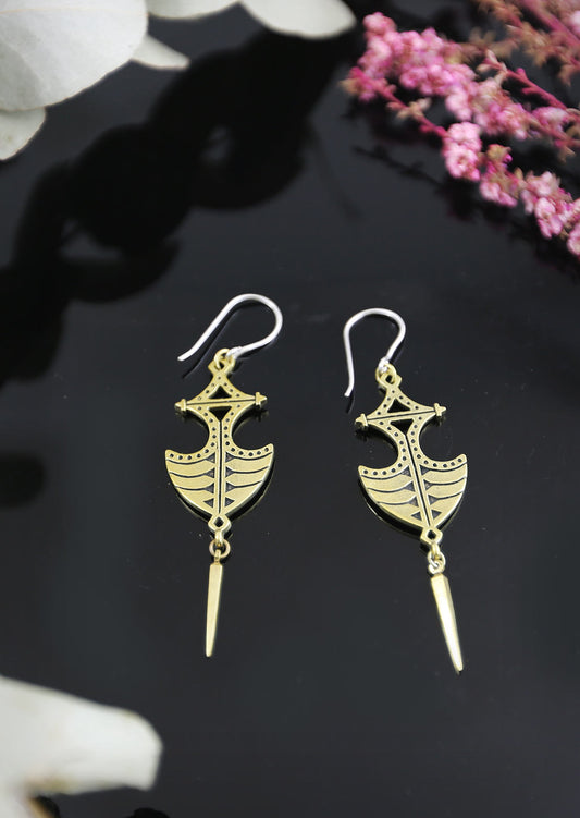 A pair of gold-colored tribal style drop earrings with sterling silver hooks, displayed against a dark background with floral elements.