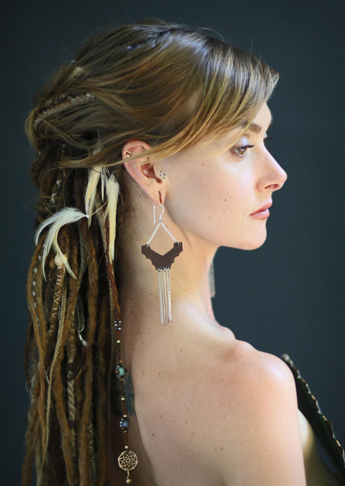 A person with a braided hairstyle wearing Ridge earrings with brass soft chains dangling down, hooks made from sterling silver, and a fabric bag.