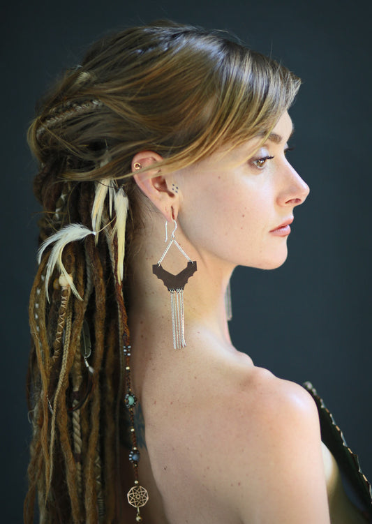 A person with a braided hairstyle wearing Ridge earrings with brass soft chains dangling down, hooks made from sterling silver, and a fabric bag.