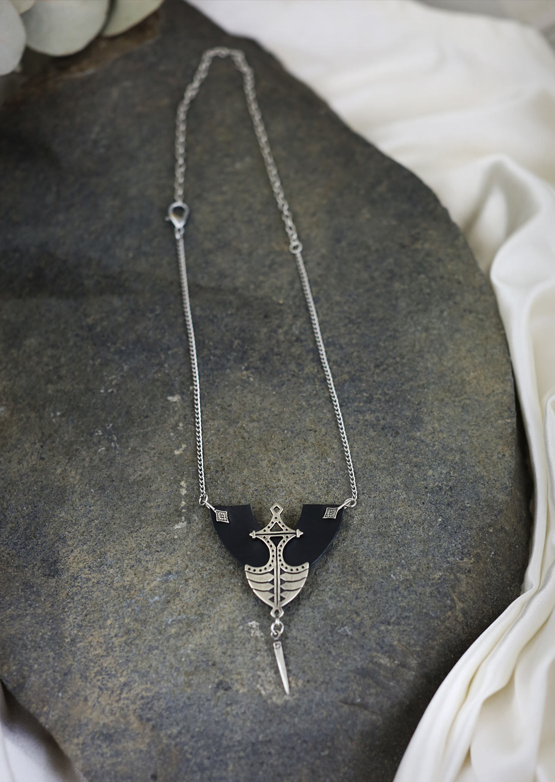 An African-inspired tribal design necklace with a leather half-spade shape and a pendulum tip, displayed on a textured background.