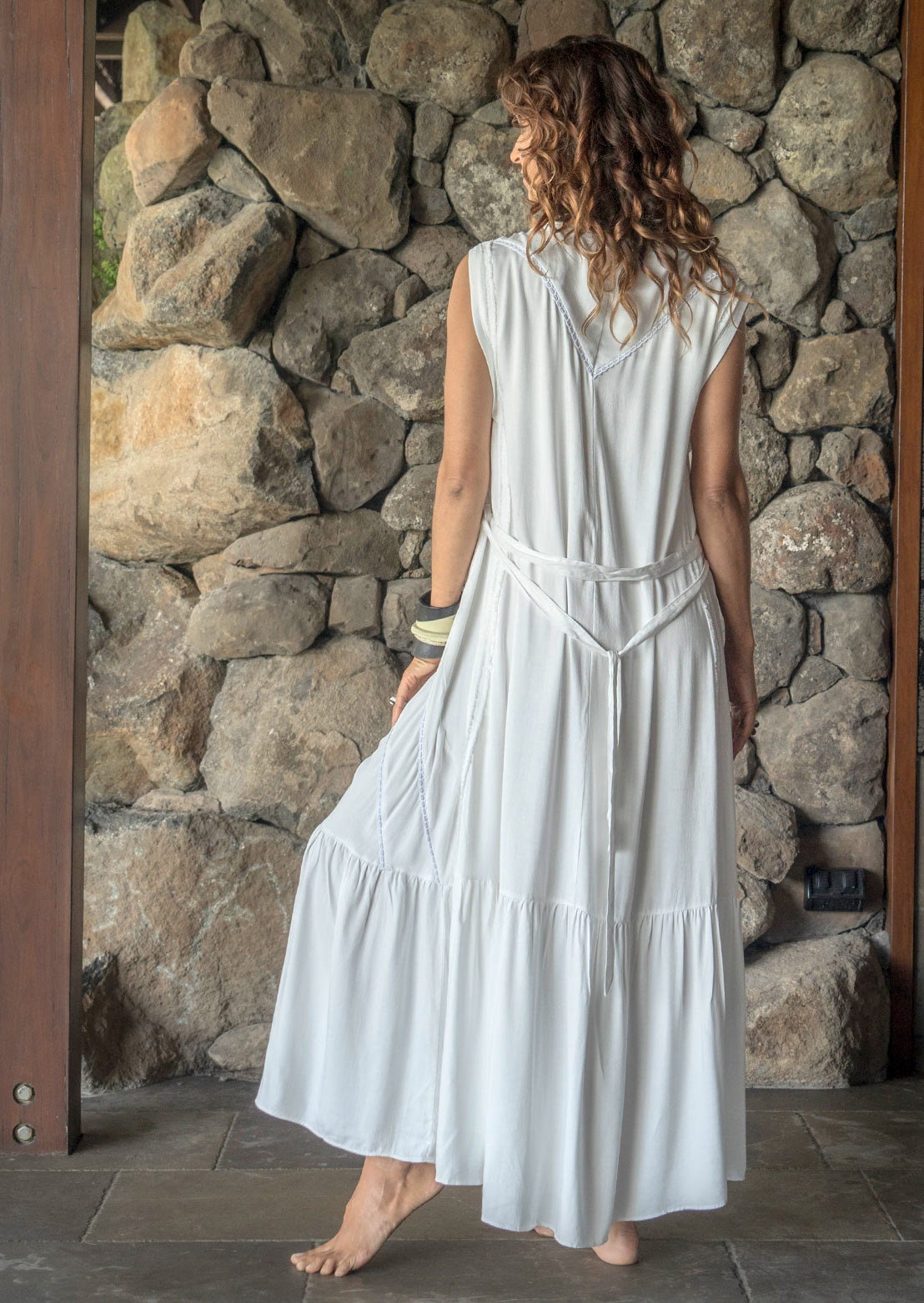 A woman standing in front of a stone wall wearing a white maxi dress with short sleeves, a V-neckline, and a flowy skirt.