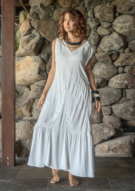 A woman standing in front of a stone wall wearing a white maxi dress with short sleeves, a V-neckline, and a flowy skirt.