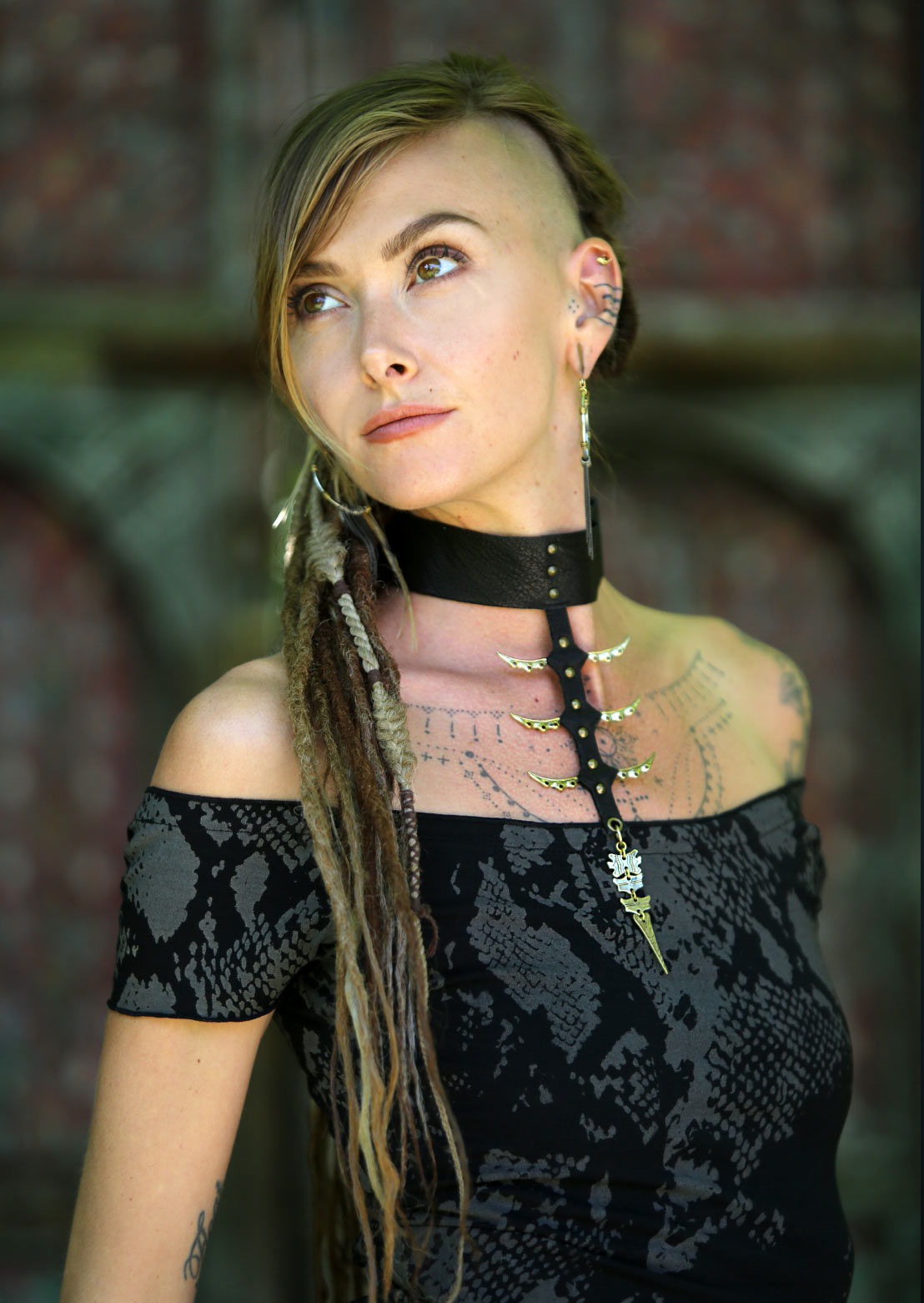A person wearing a black choker necklace with a spiked leather strap and brass/gold brass accents, standing in a dark, textured background.
