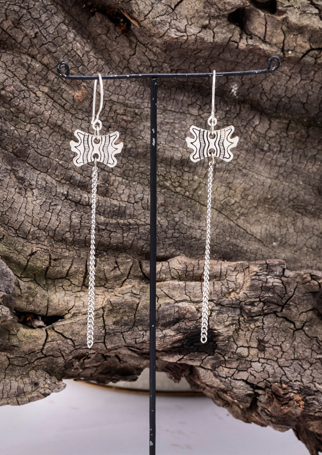 A pair of brass earrings with a geometric pattern, displayed on a black stand, set against a natural outdoor background with a wooden texture.