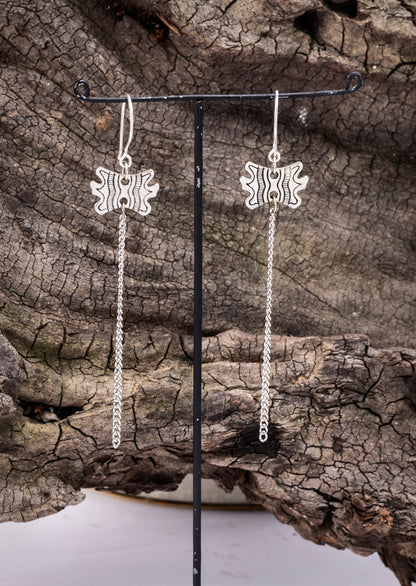 A pair of brass earrings with a geometric pattern, displayed on a black stand, set against a natural outdoor background with a wooden texture.
