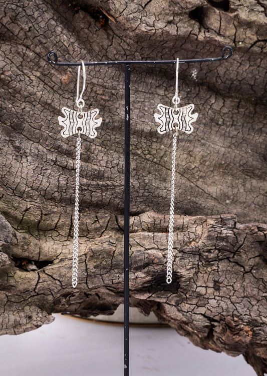 A pair of brass earrings with a geometric pattern, displayed on a black stand, set against a natural outdoor background with a wooden texture.
